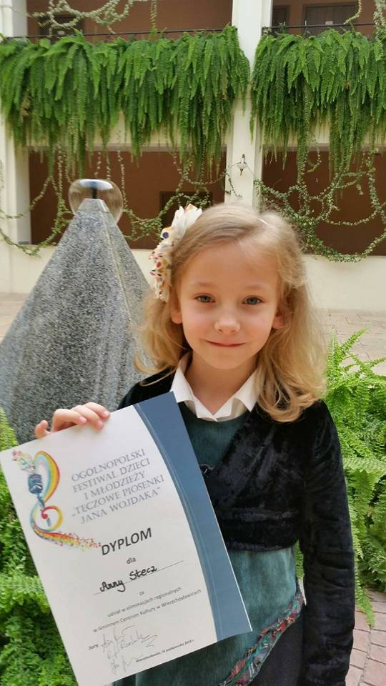 Zdjęcie przedstawia Anię Stecz, która pozuje do zdjęcia z dyplomem po eliminacjach do XVI Ogólnopolskiego Festiwalu Dzieci i Młodzieży "Tęczowe Piosenki Jana Wojdaka"