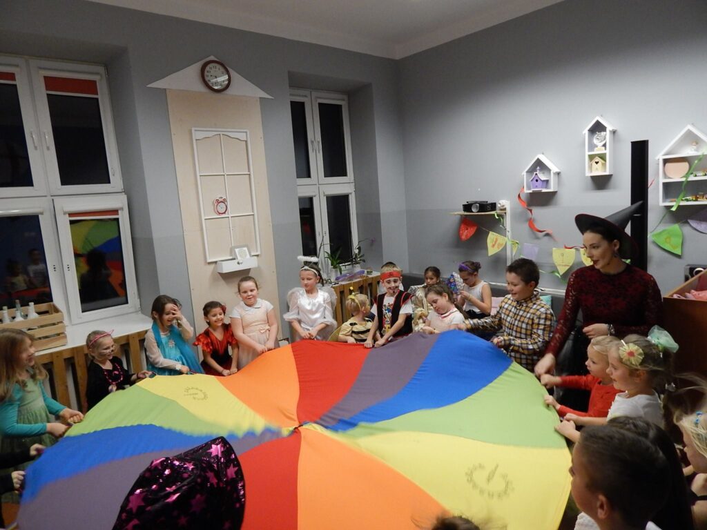 Zdjęcie przedstawia dzieci podczas zabawy Andrzejkowej trzymające kolorowe koło w pomieszczeniu z szarymi ścianami.