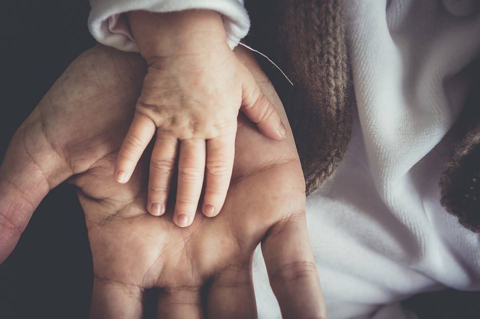 Obraz przedstawia dłoń dorosłego dotykaną przez dłoń malutkiego dziecka.