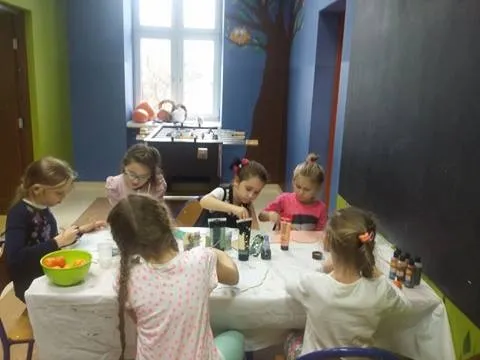 Zdjęcie przedstawia dzieci przy stole w niewielkim kolorowym pokoju, które tworzą prace plastyczne.