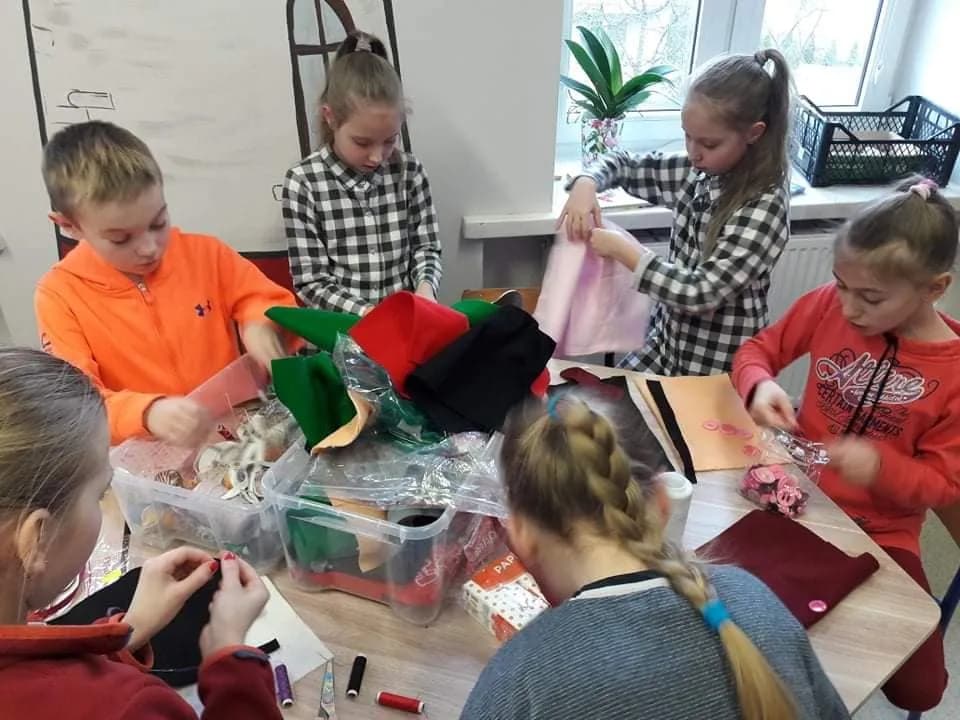 Obraz przedstawia wychowanków SPWD podczas zajęć trwających w ferie. Podczas zajęć dzieci szyją z filcu i bawełny, a kartonowe pudła zamieniają w pałace.