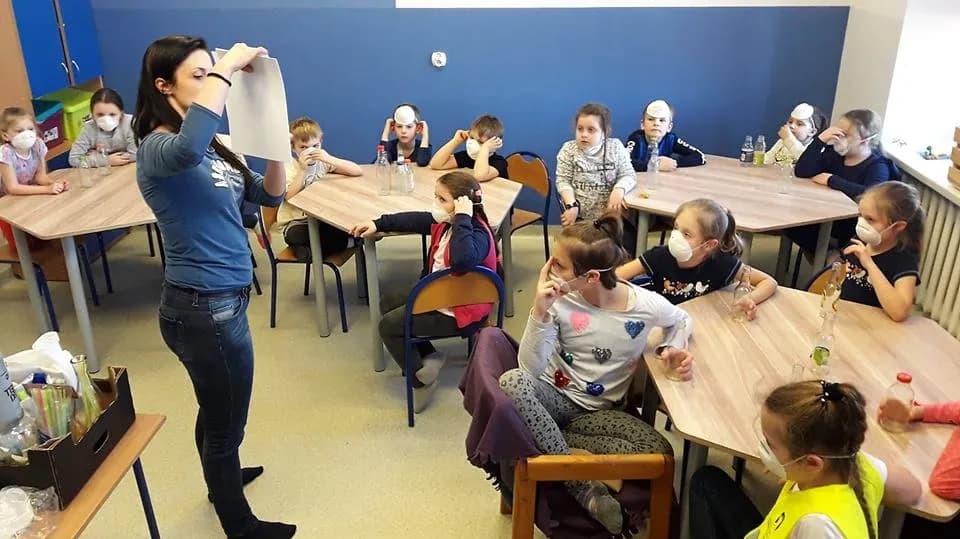 Obraz przedstawia wychowanków podczas zajęć z wykorzystaniem małego laboratorium. Na zdjęciu możemy ujrzeć dzieci siedzące przy stołach na których znajdują się przedmioty potrzebne do wykonania eksperymentów.