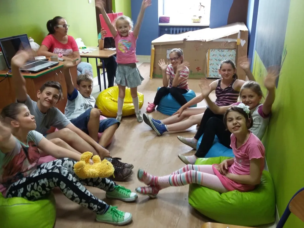 Zdjęcie przedstawia dzieci z placówki machające rękami do kamery podczas warsztatów improwizacji teatralnej w placówce.