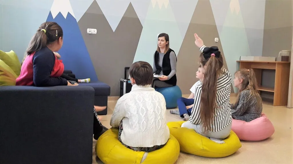 Obraz przedstawia wychowanków SPWD podczas ćwiczeń inteligencji emocjonalnej. Na zdjęciu możemy ujrzeć dzieci siedzą na poduszkach oraz sofie.