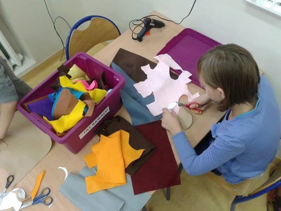 Zdjęcie przedstawia osobę wycinającą kawałek z większego materiału w kolorze białym. Obok w pudełku więcej kolorowych kawałków materiału.