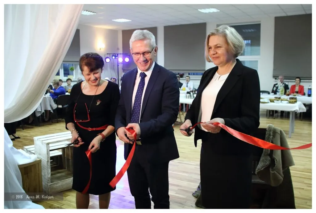 Obraz przedstawia uroczyste otwarcie Klubu Seniora, a mianowicie przecięcie czerwonej wstęgi przez burmistrza Gminy Radłów.