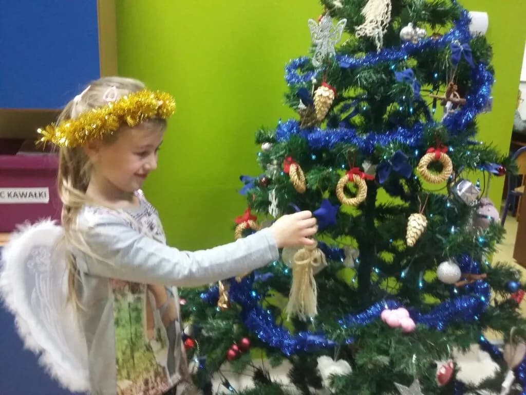 Zdjęcie przedstawia dziewczynkę w skrzydłach anioła ubierającą choinkę na tle zielonej ściany.