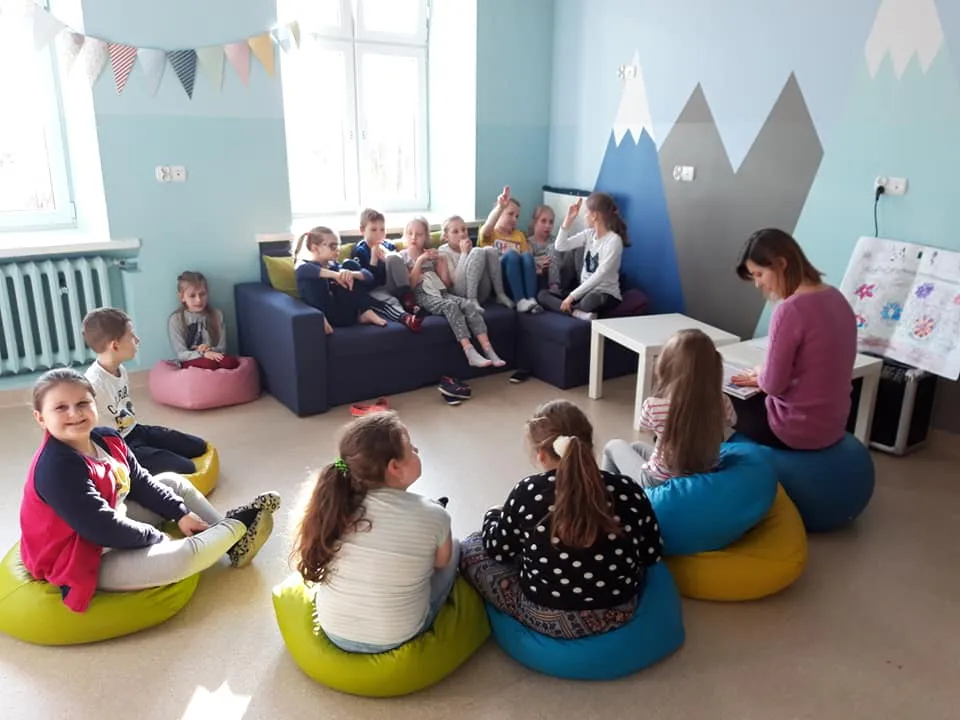 Obraz przedstawia zajęcia psychoedukacyjne. Na zdjęciu możemy ujrzeć dzieci słuchające wychowawczyni, siedzące na sofie oraz poduszkach.