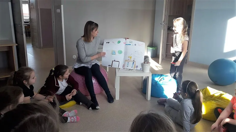 Obraz przedstawia zajęcia psychoprofilaktyczne. Na zdjęciu możemy ujrzeć dzieci z wychowawczynią, trzymająca rysunek jednej z wychowanek.