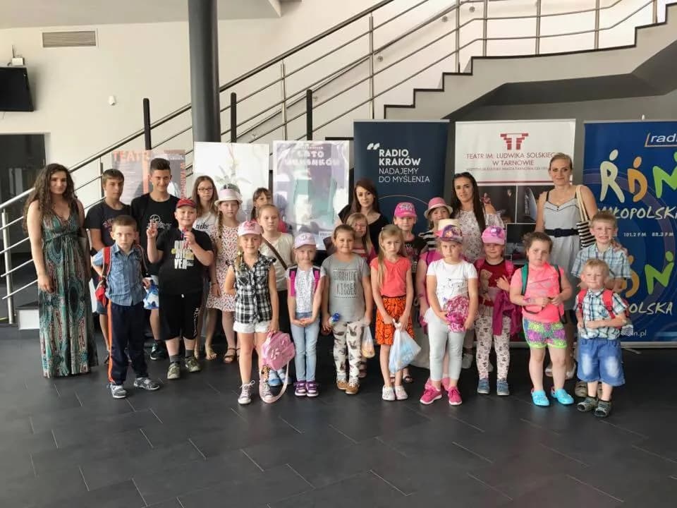Zdjęcie przedstawia dzieci oraz wychowawców placówki w teatrze. Na tle widnieją banery reklamowe oraz schody.