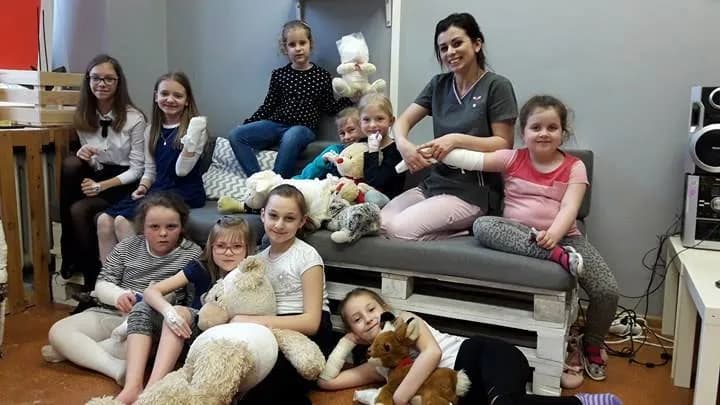 Zdjęcie przedstawia dzieci z placówki oraz osobę opiekującą się nimi siedzących na kanapie z palet oraz przed nią. Wszystko jest na tle szarej ściany.