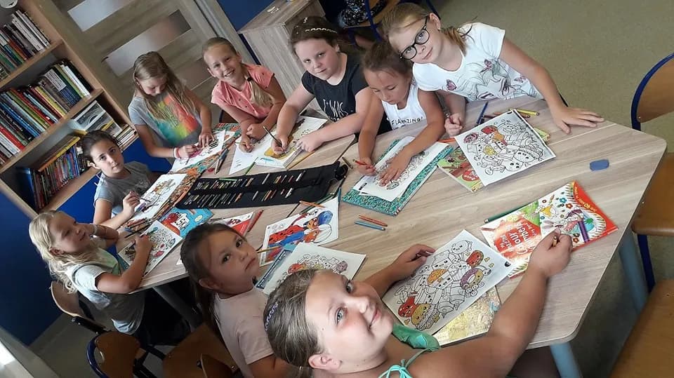 Obraz przedstawia wychowanków SPWD podczas wakacyjnych zajęć artystycznych. Na zdjęciu dzieci malują różnego rodzaju kolorowanki przy użyciu kredek.