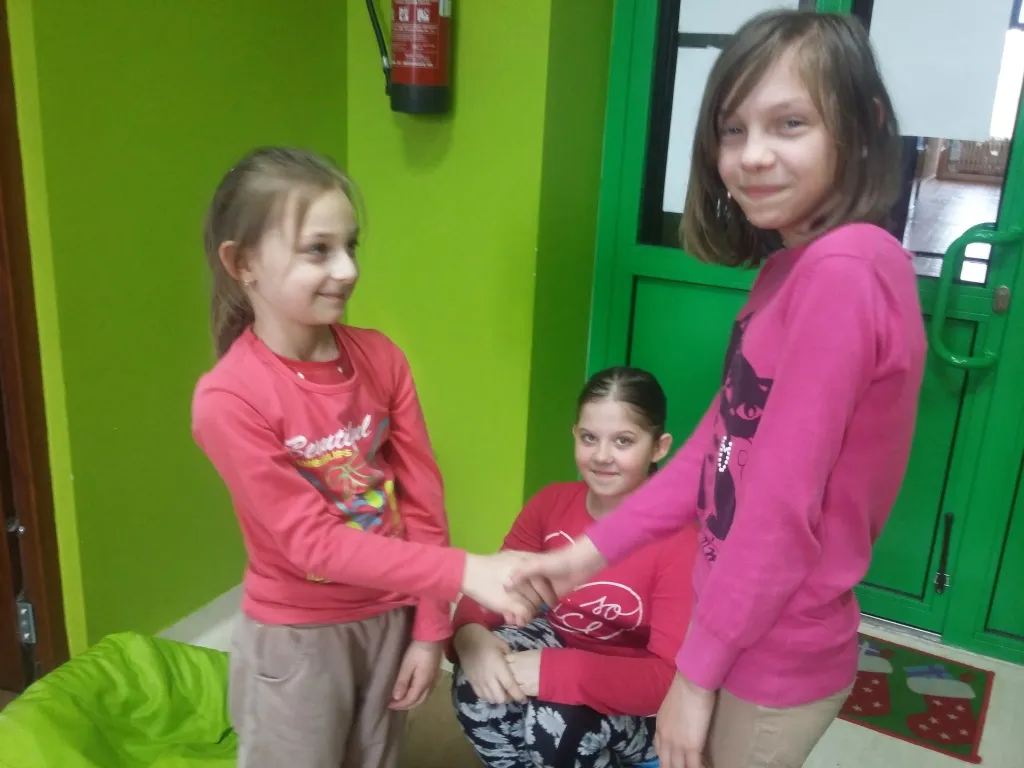 Zdjęcie przedstawia trzy dziewczynki przy wejściu, dwie z nich podają sobie ręce, natomiast trzecie siedzi w oddali.